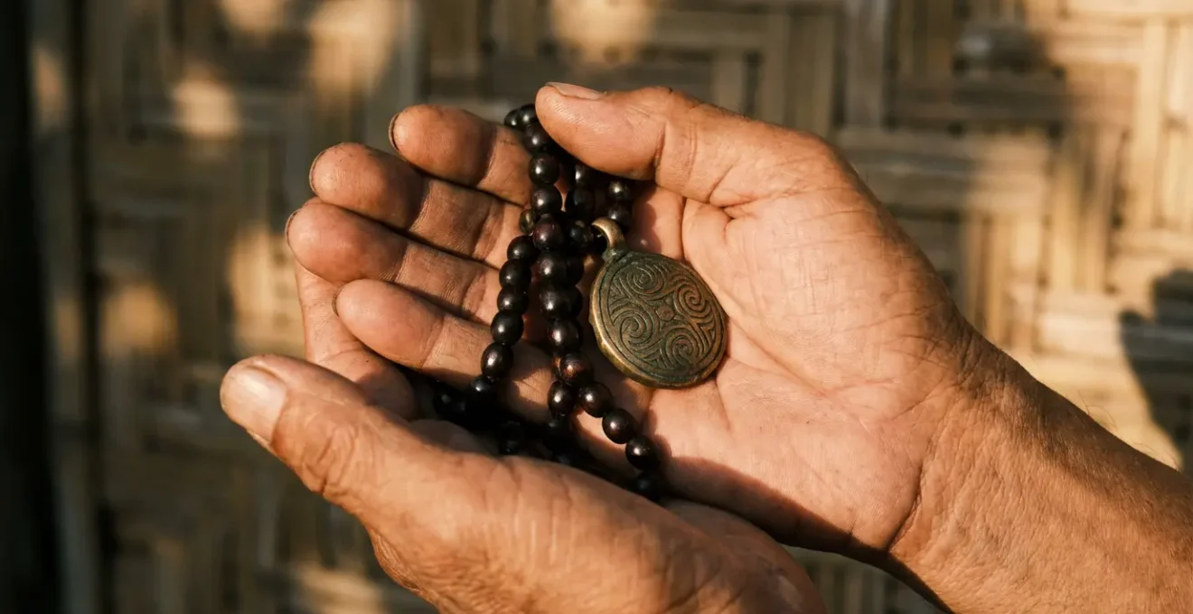 A Filipino devotee holding both a wooden rosary and traditional brass medallion amulet in weathered hands